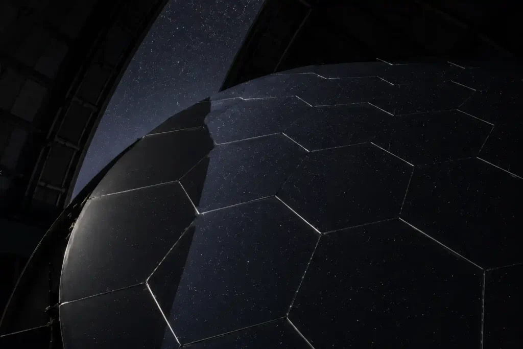 Close view of a large telescope mirror made of polished hexagonal segments inside a dark engineering facility.